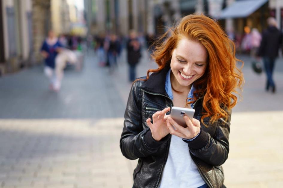 tjej står på en gågata och ler och tittar ner i sin mobil.jpg